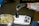 A person wearing a white lab coat and black gloves is placing cannabis buds into a black container on an electronic scale. A metal bowl filled with cannabis buds sits nearby on a dark surface.