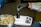 A person wearing a white lab coat and black gloves is placing cannabis buds into a black container on an electronic scale. A metal bowl filled with cannabis buds sits nearby on a dark surface.