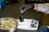 A person wearing a white lab coat and black gloves is placing cannabis buds into a black container on an electronic scale. A metal bowl filled with cannabis buds sits nearby on a dark surface.