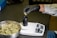 A person wearing a white lab coat and black gloves is placing cannabis buds into a black container on an electronic scale. A metal bowl filled with cannabis buds sits nearby on a dark surface.