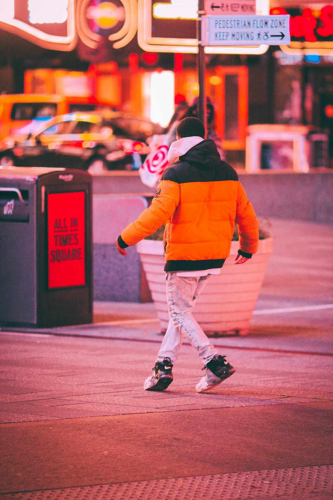 child in orange jacket and white pants standing on red and black trash bin
