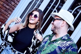 Two people are laughing and smiling, wearing stylish outfits with floral patterns. One person is in a black sleeveless top and denim shorts, accessorized with multiple rings and round sunglasses. The other person is wearing a white hat, dark sunglasses, and a vibrant shirt with green and blue leaves. They are standing in front of a brick wall and a window with decorative wrought iron.