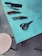 A variety of tools are placed on a light blue table, including a large cleaver, a pair of scissors, and several types of pruning shears. Nearby, a round beige stool stands on a textured concrete floor.