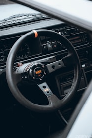 A close-up view of a sporty OMP steering wheel inside a car, with a detailed dashboard visible in the background. The steering wheel features a logo in the center with visible bolts and has a black and metallic design with an orange accent. The dashboard includes dials and switches, suggesting a classic or vintage car interior.