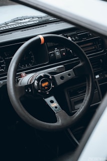 A close-up view of a sporty OMP steering wheel inside a car, with a detailed dashboard visible in the background. The steering wheel features a logo in the center with visible bolts and has a black and metallic design with an orange accent. The dashboard includes dials and switches, suggesting a classic or vintage car interior.