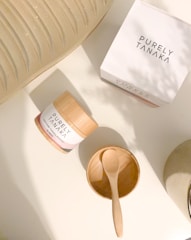 A container labeled 'PURELY TANAKA' with the text '100% Pure Tanaka Powder' is placed on a flat, light-colored surface. Nearby is an empty wooden bowl with a small wooden spoon resting inside. Another 'PURELY TANAKA' branded box is also present. Part of a textured, light-colored object is partially visible in the top left corner.