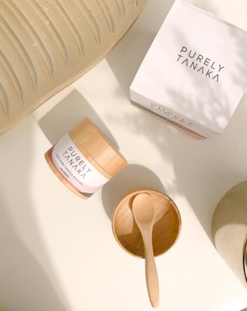A container labeled 'PURELY TANAKA' with the text '100% Pure Tanaka Powder' is placed on a flat, light-colored surface. Nearby is an empty wooden bowl with a small wooden spoon resting inside. Another 'PURELY TANAKA' branded box is also present. Part of a textured, light-colored object is partially visible in the top left corner.