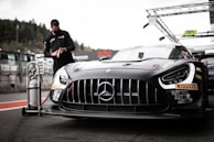 A sleek, black race car with prominent sponsor logos and a distinctive front grille is parked on a racetrack. In the background, a person wearing a mask and dark clothing is interacting with a tall gas tank on wheels. The setting appears to be a pit lane, with various equipment and structures visible.