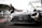 A sleek, black race car with prominent sponsor logos and a distinctive front grille is parked on a racetrack. In the background, a person wearing a mask and dark clothing is interacting with a tall gas tank on wheels. The setting appears to be a pit lane, with various equipment and structures visible.