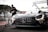 A sleek, black race car with prominent sponsor logos and a distinctive front grille is parked on a racetrack. In the background, a person wearing a mask and dark clothing is interacting with a tall gas tank on wheels. The setting appears to be a pit lane, with various equipment and structures visible.