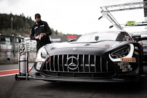 A sleek, black race car with prominent sponsor logos and a distinctive front grille is parked on a racetrack. In the background, a person wearing a mask and dark clothing is interacting with a tall gas tank on wheels. The setting appears to be a pit lane, with various equipment and structures visible.