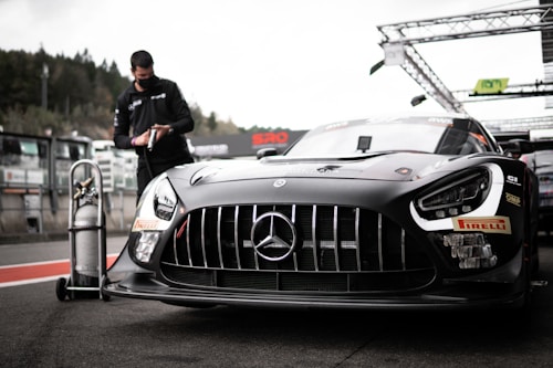 A sleek, black race car with prominent sponsor logos and a distinctive front grille is parked on a racetrack. In the background, a person wearing a mask and dark clothing is interacting with a tall gas tank on wheels. The setting appears to be a pit lane, with various equipment and structures visible.