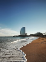 A sandy beach stretches towards a skyline with gentle waves lapping at the shore. In the distance, a distinctive sail-shaped skyscraper stands tall against a clear blue sky.