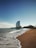 A sandy beach stretches towards a skyline with gentle waves lapping at the shore. In the distance, a distinctive sail-shaped skyscraper stands tall against a clear blue sky.