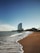 A sandy beach stretches towards a skyline with gentle waves lapping at the shore. In the distance, a distinctive sail-shaped skyscraper stands tall against a clear blue sky.