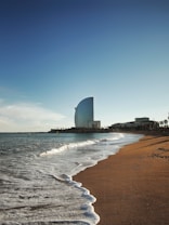 A sandy beach stretches towards a skyline with gentle waves lapping at the shore. In the distance, a distinctive sail-shaped skyscraper stands tall against a clear blue sky.