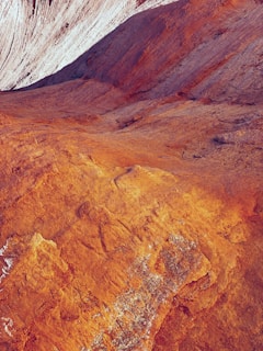 The image depicts a rugged, textured landscape with vivid orange and red hues, suggesting a terrain of eroded rock or desert. The surface appears uneven with visible striations and layers, possibly indicating geological formations.