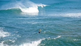 A group of surfers is scattered across a vast ocean, with powerful waves rolling in. Two surfers are paddling on their boards in the foreground, moving towards a large, cresting wave. The scene is bathed in bright, natural light, highlighting the vibrant blue and turquoise colors of the water.