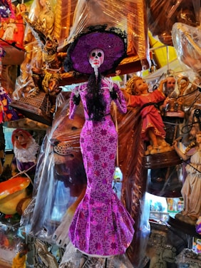A skeletal figurine dressed in an ornate purple gown and a large matching hat, surrounded by various decorative items including religious statues and colorful figures. The intricate pattern on the dress and hat adds vibrancy to the scene, and the figurine's skull is painted with exaggerated features.