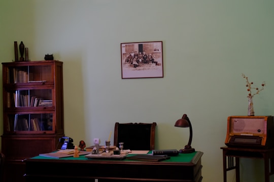 A vintage office setting with a wooden desk topped with various items including papers, a lamp, and a telephone. A bookshelf filled with books stands on the left, and a retro radio is placed on a small table to the right. A framed photograph is mounted on the pale green wall above the desk.