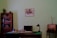 A vintage office setting with a wooden desk topped with various items including papers, a lamp, and a telephone. A bookshelf filled with books stands on the left, and a retro radio is placed on a small table to the right. A framed photograph is mounted on the pale green wall above the desk.