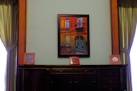 A framed painting hangs on a wall, depicting a vivid architectural scene with a door and windows in bold red and blue hues. The wall is flanked by dark wooden paneling and green draped curtains. On a dark, polished wooden cabinet below the painting are three framed photographs, evenly spaced along the surface.