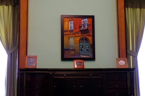 A framed painting hangs on a wall, depicting a vivid architectural scene with a door and windows in bold red and blue hues. The wall is flanked by dark wooden paneling and green draped curtains. On a dark, polished wooden cabinet below the painting are three framed photographs, evenly spaced along the surface.