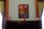 A framed painting hangs on a wall, depicting a vivid architectural scene with a door and windows in bold red and blue hues. The wall is flanked by dark wooden paneling and green draped curtains. On a dark, polished wooden cabinet below the painting are three framed photographs, evenly spaced along the surface.