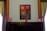 A framed painting hangs on a wall, depicting a vivid architectural scene with a door and windows in bold red and blue hues. The wall is flanked by dark wooden paneling and green draped curtains. On a dark, polished wooden cabinet below the painting are three framed photographs, evenly spaced along the surface.