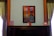 A framed painting hangs on a wall, depicting a vivid architectural scene with a door and windows in bold red and blue hues. The wall is flanked by dark wooden paneling and green draped curtains. On a dark, polished wooden cabinet below the painting are three framed photographs, evenly spaced along the surface.
