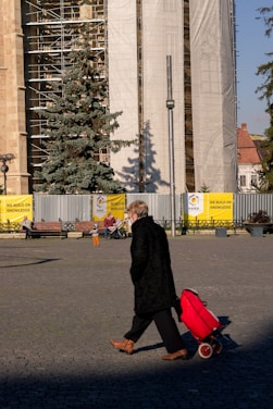 A person pulling a red trolley bag walks across a cobblestone square in front of a large building under construction with scaffolding and protective coverings. There are several benches along the building with people sitting and a large evergreen tree next to the construction site. Banners with the text 'We Build On Knowledge' are placed in front of the construction site.