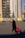 A person pulling a red trolley bag walks across a cobblestone square in front of a large building under construction with scaffolding and protective coverings. There are several benches along the building with people sitting and a large evergreen tree next to the construction site. Banners with the text 'We Build On Knowledge' are placed in front of the construction site.