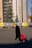 A person pulling a red trolley bag walks across a cobblestone square in front of a large building under construction with scaffolding and protective coverings. There are several benches along the building with people sitting and a large evergreen tree next to the construction site. Banners with the text 'We Build On Knowledge' are placed in front of the construction site.