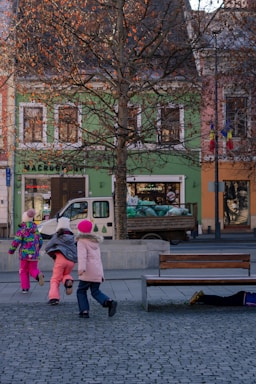 A group of children are wearing colorful winter clothes, running energetically through a paved town square. Trees with sparse leaves are in the foreground, and a row of vibrant buildings with a shop and a parked truck are in the background. One child appears to be playing or hiding under a wooden bench.