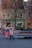 A group of children are wearing colorful winter clothes, running energetically through a paved town square. Trees with sparse leaves are in the foreground, and a row of vibrant buildings with a shop and a parked truck are in the background. One child appears to be playing or hiding under a wooden bench.