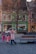 A group of children are wearing colorful winter clothes, running energetically through a paved town square. Trees with sparse leaves are in the foreground, and a row of vibrant buildings with a shop and a parked truck are in the background. One child appears to be playing or hiding under a wooden bench.