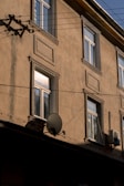 A beige exterior wall of a building with several windows and architectural moldings. There is a satellite dish attached to the wall, along with an air conditioning unit. Power lines run across the front of the building.