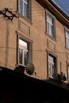 A beige exterior wall of a building with several windows and architectural moldings. There is a satellite dish attached to the wall, along with an air conditioning unit. Power lines run across the front of the building.