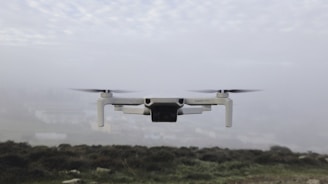 A small drone hovers in the air against a foggy backdrop. The landscape below is blurred and barely visible, suggesting a cloudy or misty day. The drone's propellers are spinning, and it appears to be of a light color, possibly white or light grey.