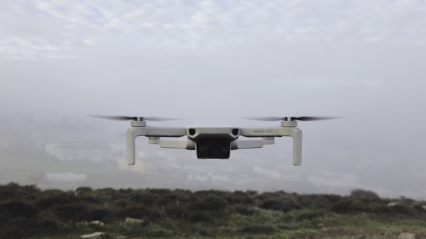 A small drone hovers in the air against a foggy backdrop. The landscape below is blurred and barely visible, suggesting a cloudy or misty day. The drone's propellers are spinning, and it appears to be of a light color, possibly white or light grey.