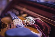 A close-up of a violin placed inside its case with a small, whimsical figurine resembling a fictional character nestled beside it on velvet fabric. The image captures intricate details of the violin's strings and bridge, set against a warm and soft-focus background.