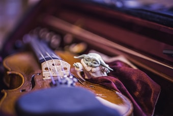 A close-up of a violin placed inside its case with a small, whimsical figurine resembling a fictional character nestled beside it on velvet fabric. The image captures intricate details of the violin's strings and bridge, set against a warm and soft-focus background.