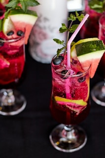 A vibrant, chilled beverage in a tall glass garnished with slices of watermelon and lime. The drink is a deep red color and features fresh blueberries and mint leaves, with a pink straw for sipping. Other similar glasses are visible around it, placed on a dark surface.