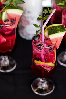 A vibrant, chilled beverage in a tall glass garnished with slices of watermelon and lime. The drink is a deep red color and features fresh blueberries and mint leaves, with a pink straw for sipping. Other similar glasses are visible around it, placed on a dark surface.
