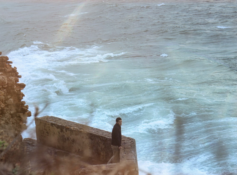 homem na jaqueta preta sentado na parede de concreto marrom perto das ondas do mar durante o dia