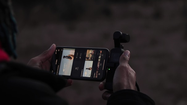 A person holds a smartphone in one hand and a small camera gimbal in the other. The smartphone screen displays an app with preview images, possibly related to video or photography.