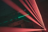 A series of bright red laser beams extend diagonally across a dark, industrial space. The background is a dimly lit area with hints of green light reflected on a textured surface, possibly a metal structure.