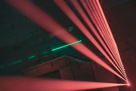 A series of bright red laser beams extend diagonally across a dark, industrial space. The background is a dimly lit area with hints of green light reflected on a textured surface, possibly a metal structure.