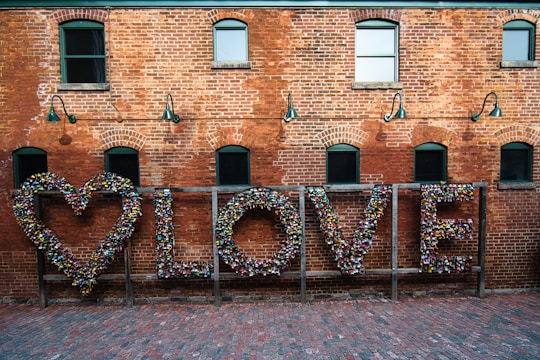 A red brick wall with several arched windows and green framed lamps. In front of the wall, a wooden frame holds a heart shape and the word 'LOVE' made out of numerous colorful locks.