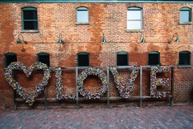 A red brick wall with several arched windows and green framed lamps. In front of the wall, a wooden frame holds a heart shape and the word 'LOVE' made out of numerous colorful locks.